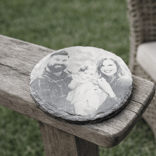 A Custom Laser Engraved Slate Coaster featuring a black-and-white photo of a man, woman, and baby, displayed on an outdoor wooden bench with grass and a wicker chair in the background.