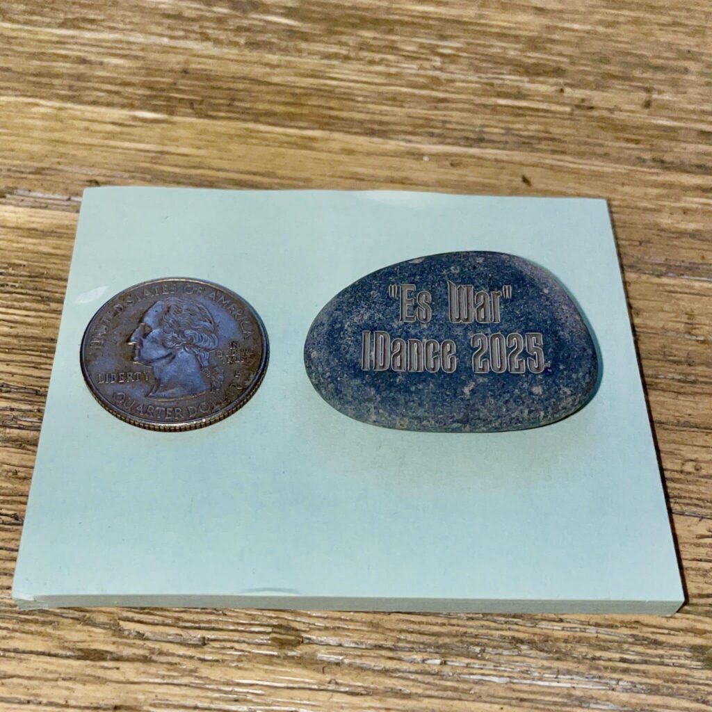 A 1/2" Mini Engraved River Stone with "Es War iDance 2025" and a U.S. quarter sit on a blue sticky note atop a wooden surface.