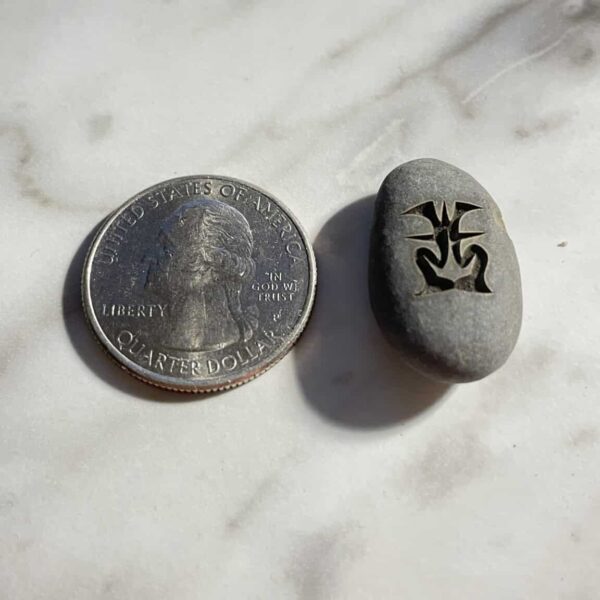 A U.S. quarter sits beside a 1/2" Mini Engraved River Stone featuring a black abstract symbol, both displayed on a marble surface.