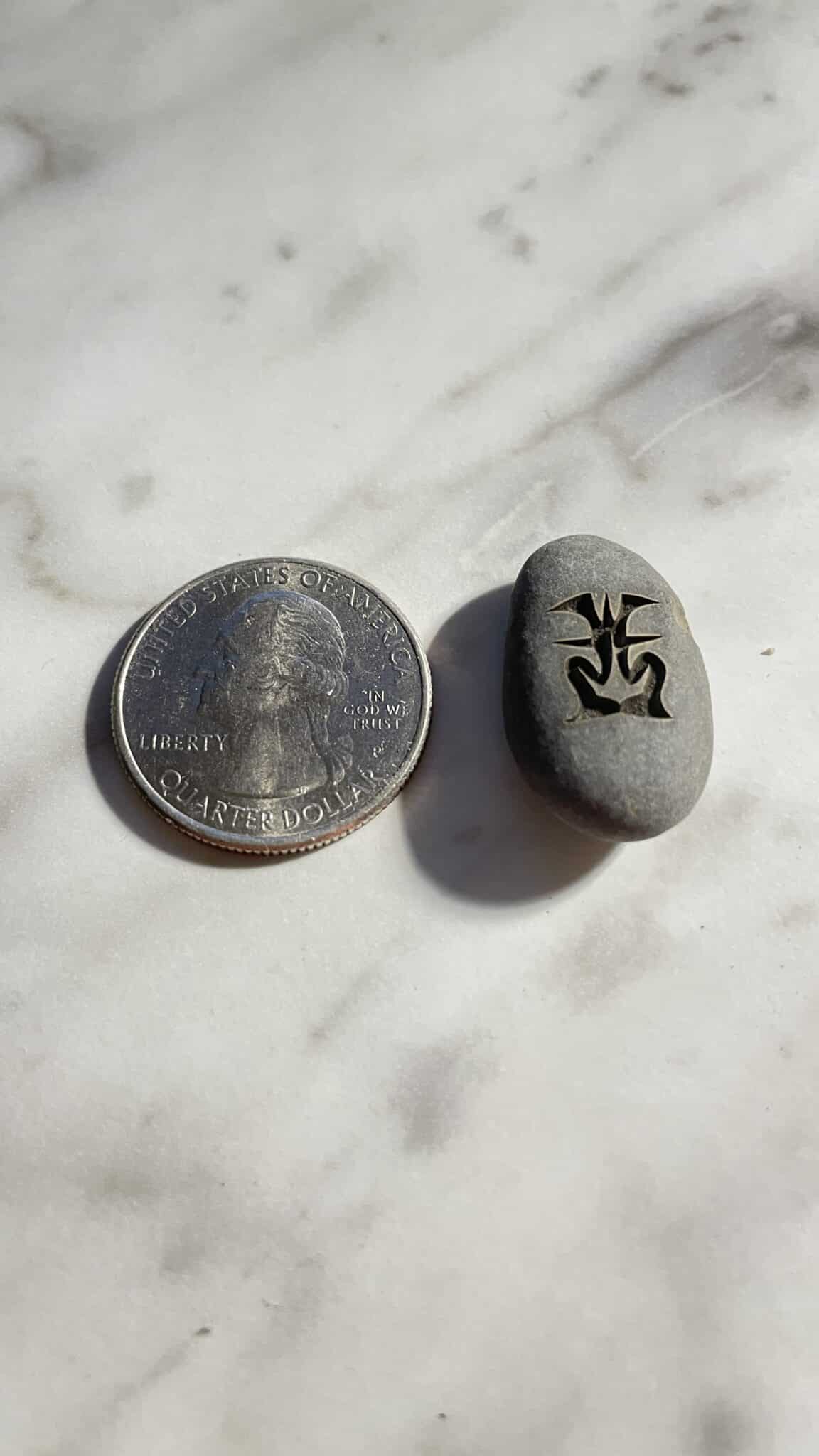 A U.S. quarter sits beside a 1/2" Mini Engraved River Stone featuring a black abstract symbol, both displayed on a marble surface.