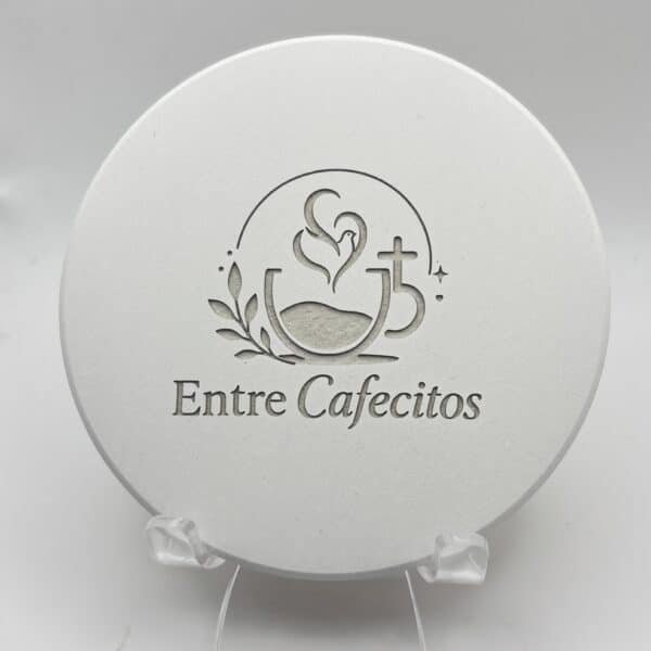 A round white MagicStone Engraved Tile Coaster featuring the words “Entre Cafecitos” below an illustration of a coffee cup with heart-shaped steam, a small cross, and leaves, displayed on a clear stand.
