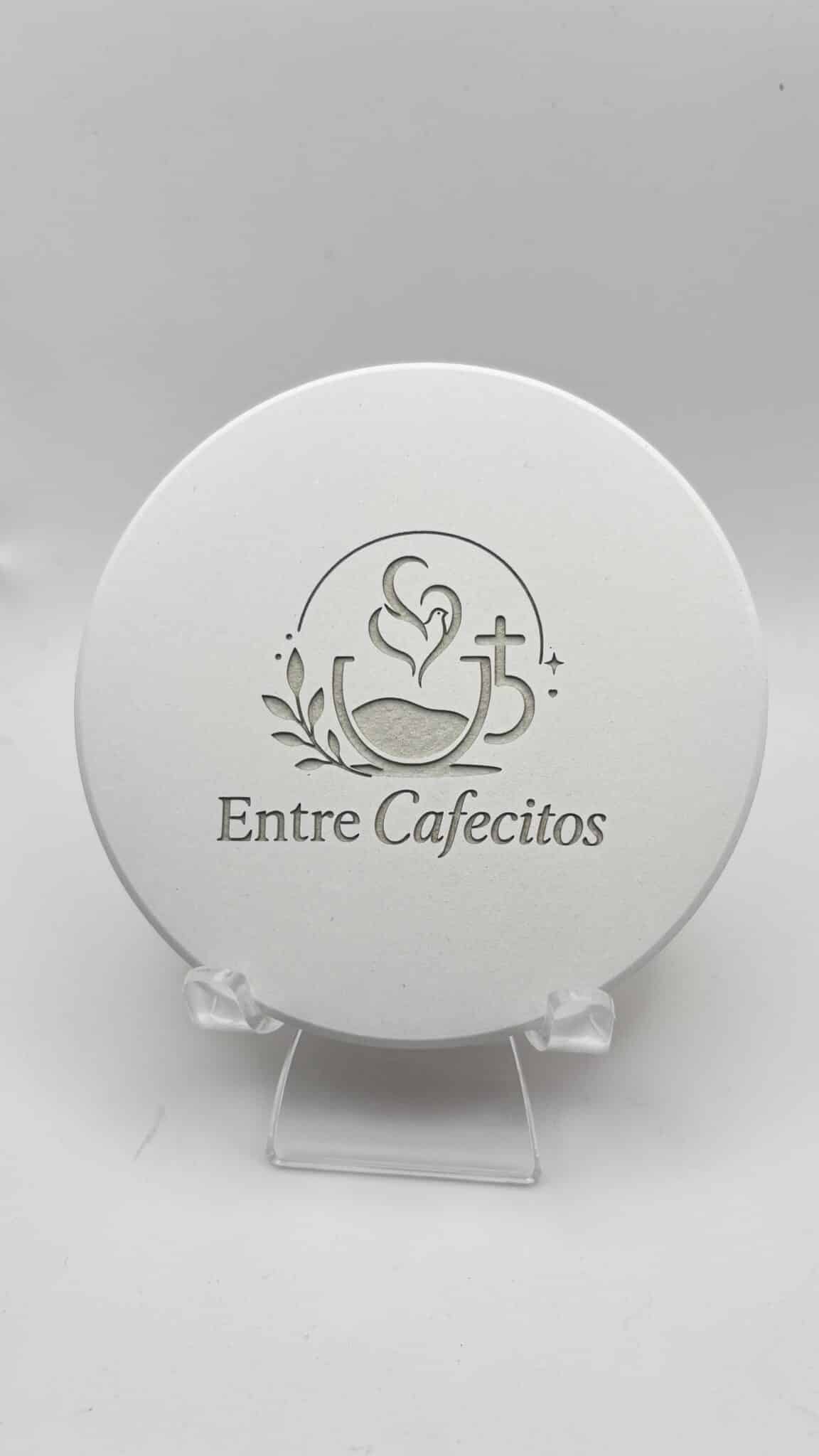 A round white MagicStone Engraved Tile Coaster featuring the words “Entre Cafecitos” below an illustration of a coffee cup with heart-shaped steam, a small cross, and leaves, displayed on a clear stand.