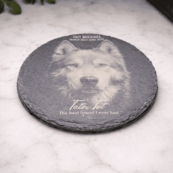 A Custom Laser Engraved Slate Coaster featuring a wolf's face and the inscription "Tait Michael - The best friend I ever had" rests on marble, with greenery nearby.