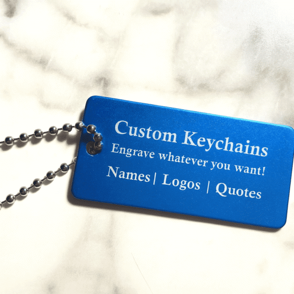A blue Metal Keychain with Optional Chain, engraved with "Custom Keychains." Personalize with names, logos, or quotes. Shown on a white marble surface.