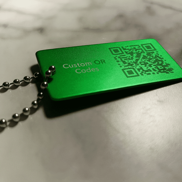 A green Metal Keychain with Optional Chain, engraved with a QR code and "Custom QR Codes," is shown resting on a marble surface.
