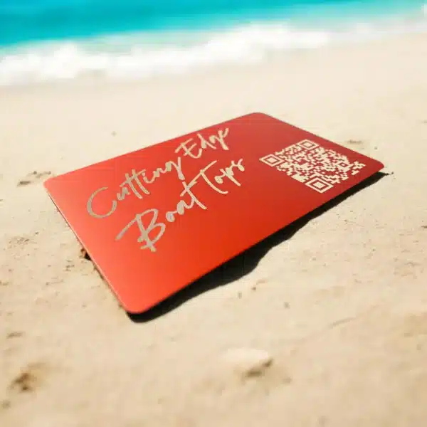 A .42mm Engraved Aluminum Business Card featuring “Cutting Edge Boat Tours” and a QR code lies on the sand, with ocean waves softly blurred in the background.