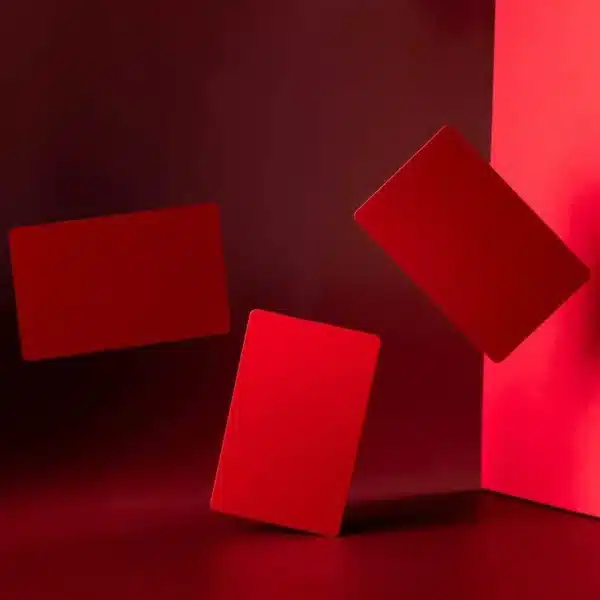 Three .8mm Engraved Aluminum Business Cards are suspended in midair against a deep red background. One card leans against a vertical red surface on the right side of the image.