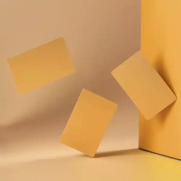 Three .8mm Engraved Aluminum Business Cards in golden-yellow float in a minimalist setting with a warm beige background, one card leaning against a vertical surface for a modern, abstract look.
