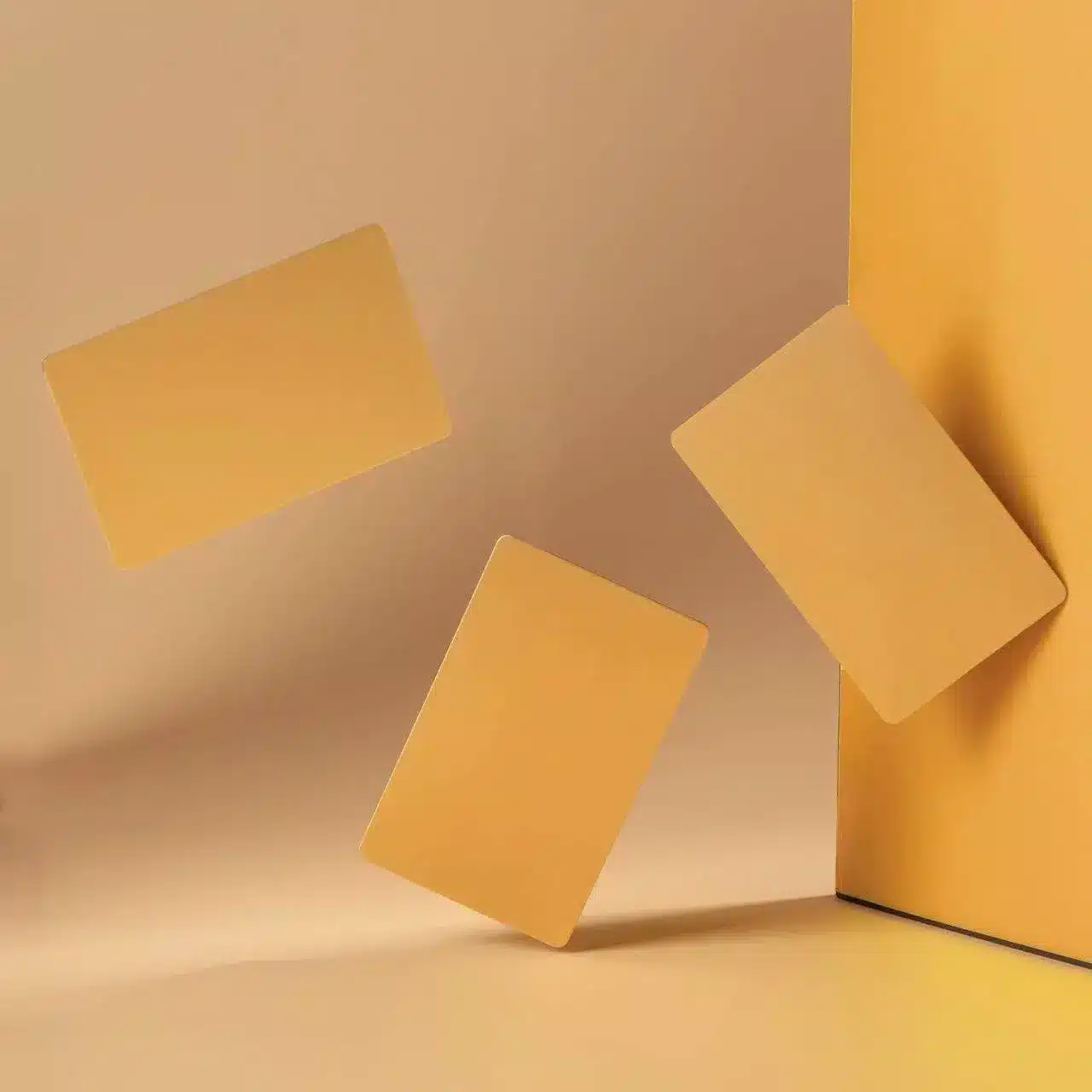 Three .8mm Engraved Aluminum Business Cards in golden-yellow float in a minimalist setting with a warm beige background, one card leaning against a vertical surface for a modern, abstract look.