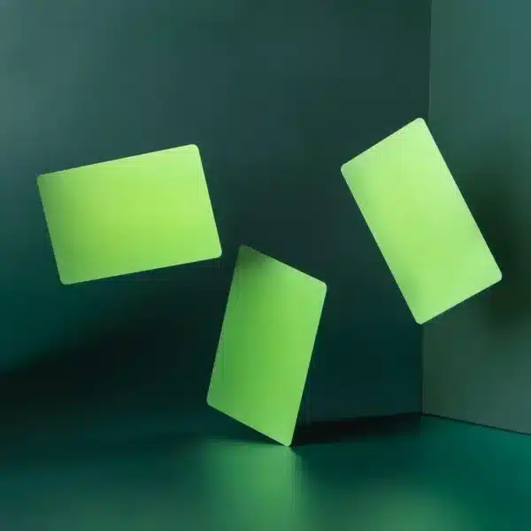 Three .8mm Engraved Aluminum Business Cards in neon green float and tilt at various angles against a dark green background and floor, with two walls forming a corner.