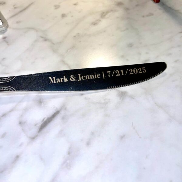 A close-up of an Engraved Stainless Steel Butter Knife with "Mark & Jennie | 7/21/2025" on its blade, resting on a white marble surface.