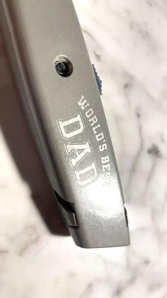 An Engraved Safety Utility Knife rests on a marble surface, with the words "WORLDS BEST DAD" engraved on its side.