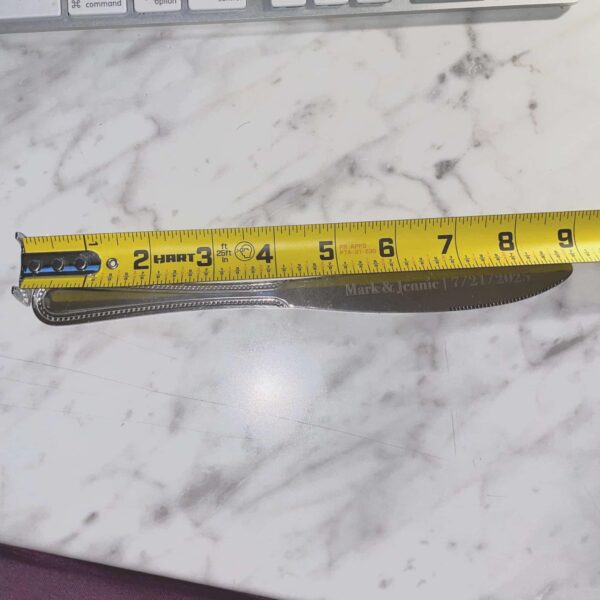 An Engraved Stainless Steel Butter Knife with "Mark & Jennie 7-27-12" on the blade is measured at about 8 inches long by a yellow tape measure on a white marble surface.