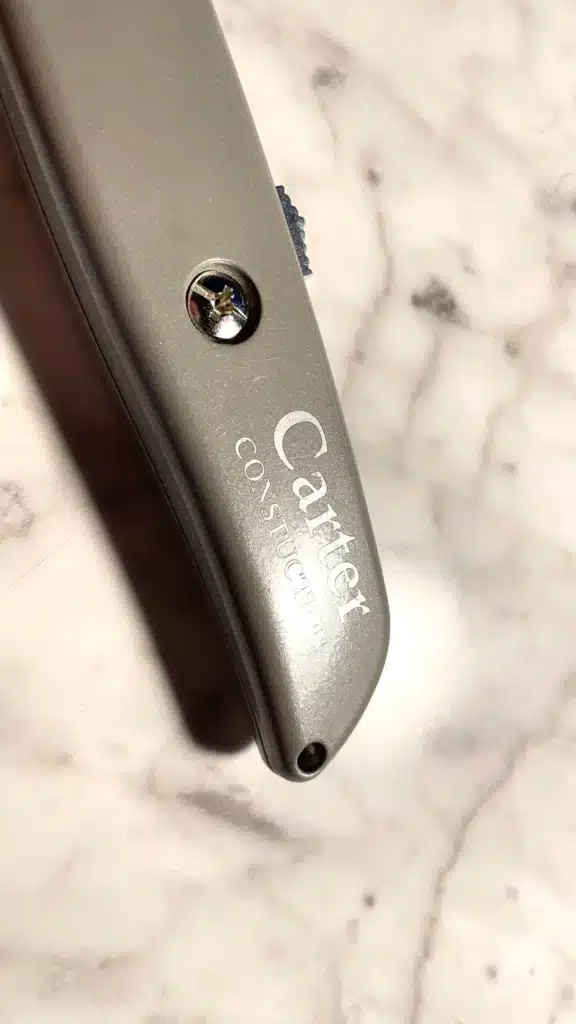 A close-up of the Engraved Safety Utility Knife, featuring the Cartier CONSTRUCTION text on silver metal atop a marble surface, with a visible screw and a small textured blue section.