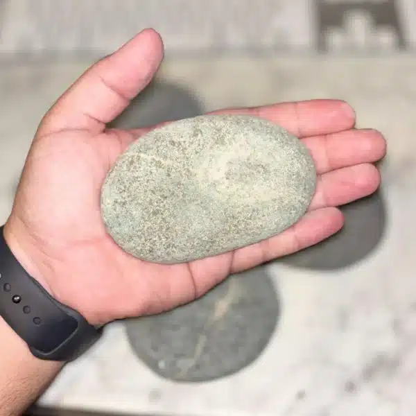A person holds a 1-5" Aged Rustic Engraved River Rock in their hand, with more similar stones blurred in the background on a light surface. The person is wearing a dark wristband.