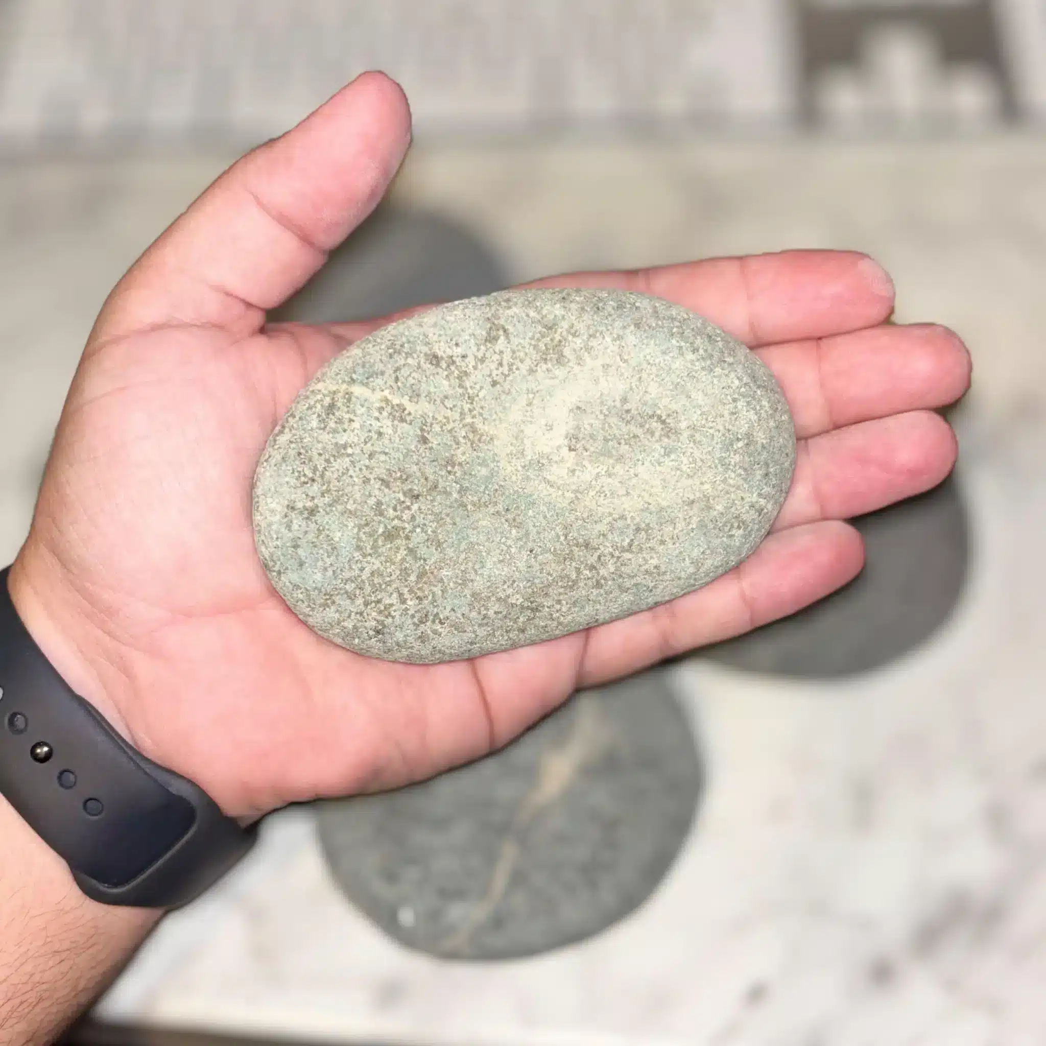 A person holds a 1-5" Aged Rustic Engraved River Rock in their hand, with more similar stones blurred in the background on a light surface. The person is wearing a dark wristband.