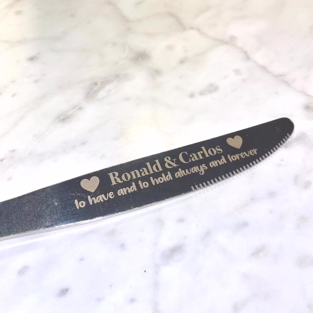 An Engraved Stainless Steel Butter Knife featuring heart motifs and the inscription "Ronald & Carlos ♥ to have and to hold always and forever," displayed on a white marble surface.