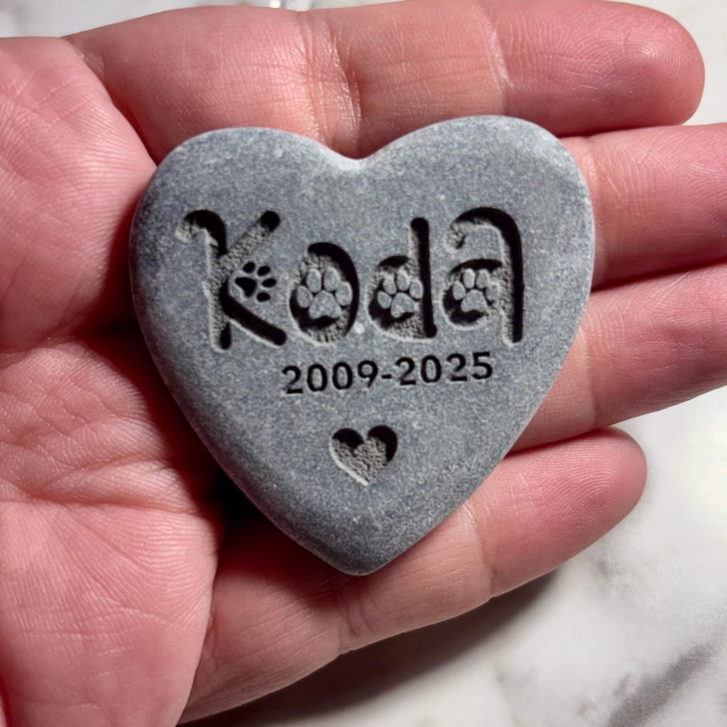 A hand holds the Engraved Heart Pet Memorial Stone – Personalized Pet Remembrance Keepsake, in gray and heart-shaped, with "Koda" (paw prints for the O), years 2009-2025, and a small engraved heart at the bottom.