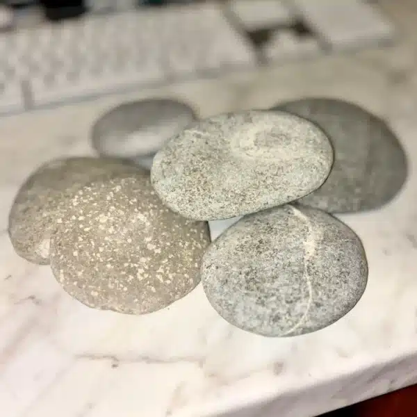 Several 1-5" Aged Rustic Engraved River Rocks, smooth and light-colored, are arranged on a marble surface, with a blurred computer keyboard and monitor in the background.