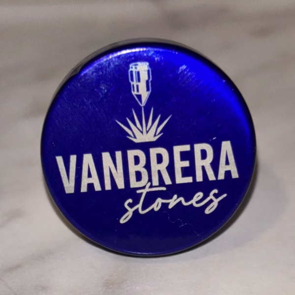 A round, metallic blue Engraved Aluminum Wine Bottle Stopper featuring white "VANBRERA stones" text below a stylized plant and vertical object graphic, set on a light marble background.