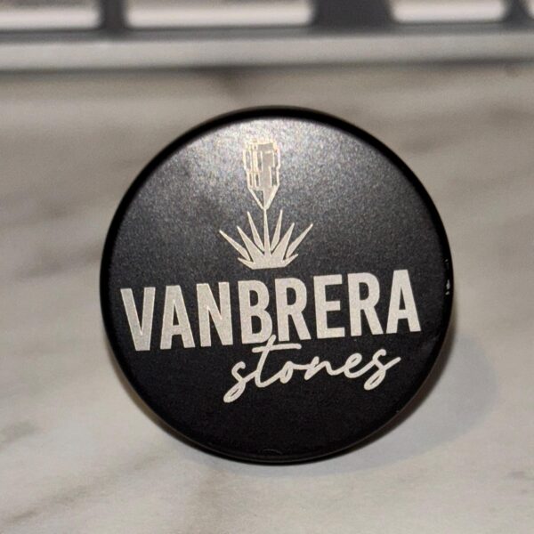A black circular Engraved Aluminum Wine Bottle Stopper lid with silver "VANBRERA stones" text and an agave piña graphic on a marble surface.