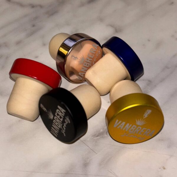 Four Engraved Aluminum Wine Bottle Stoppers with colored tops—red, gold, black, and blue—are arranged on a white marble surface.