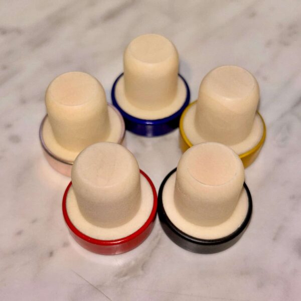 Five Engraved Aluminum Wine Bottle Stoppers with ivory cylindrical tops and bases in red, black, yellow, blue, and pink are arranged in a circle on a marble surface.