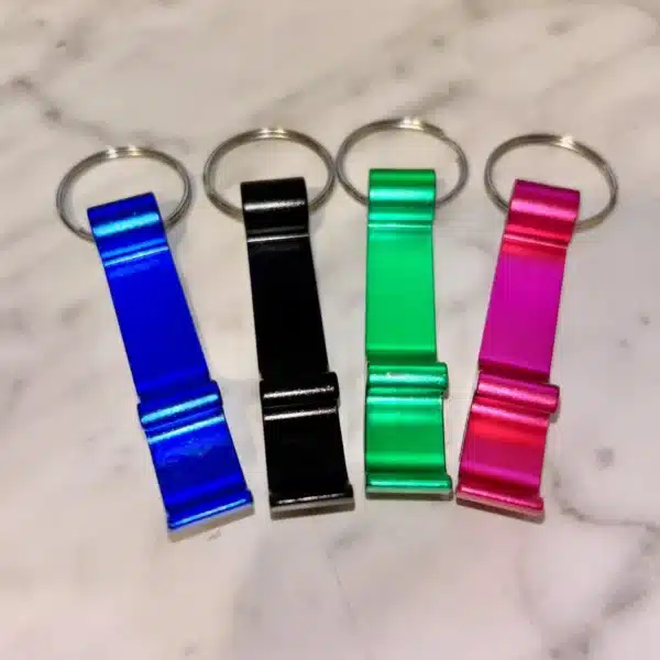 Four Custom Engraved Bottle Opener Keychains in blue, black, green, and pink are lined up side by side on a light marble surface.