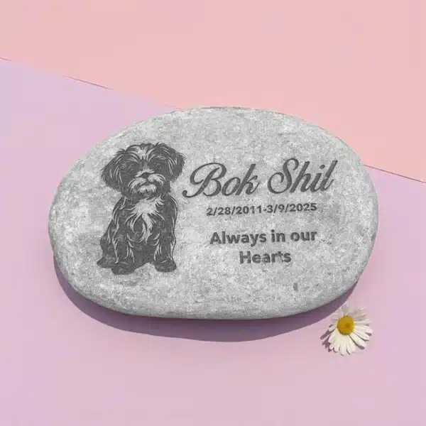 A 2–4.5" Tiny Tributes Custom Engraved Pet Memorial Stone features a small dog illustration, the name Bok Shil, the dates 2/28/2011–3/9/2025, "Always in our Hearts," and a white daisy on a pink-purple background.