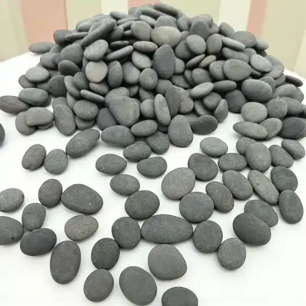 A pile of 1/2" Mini Engraved River Stones, smooth and oval-shaped in grey, is stacked on a white surface with more stones scattered in front. The background features pastel vertical stripes.