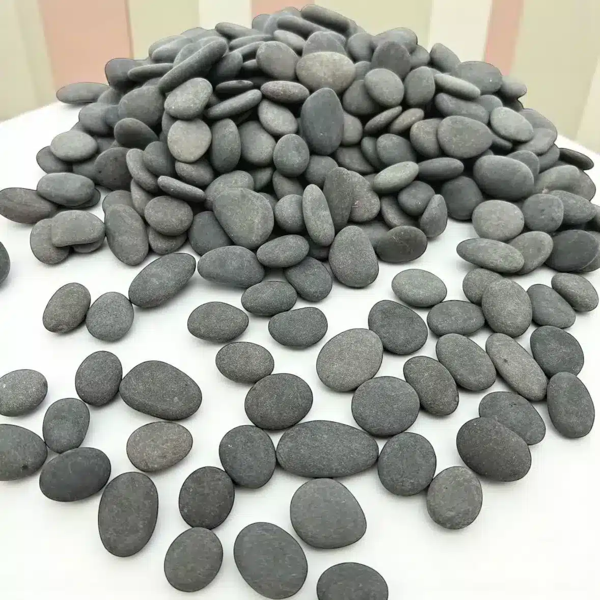 A pile of 1/2" Mini Engraved River Stones, smooth and oval-shaped in grey, is stacked on a white surface with more stones scattered in front. The background features pastel vertical stripes.
