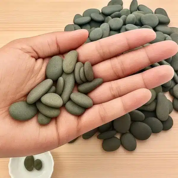 A hand holds several 1/2" Mini Engraved River Stones, smooth and oval-shaped, with more of these dark green stones scattered on a wooden surface in the background.