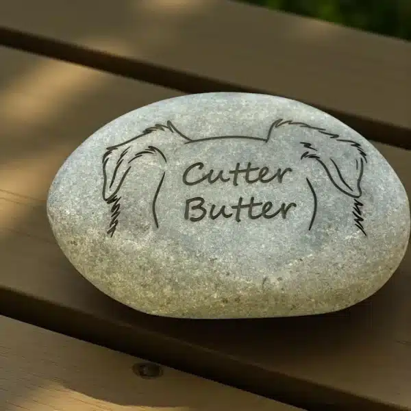 A smooth gray 2–4.5" Tiny Tributes Custom Engraved Pet Memorial Stone rests on wood, featuring a sketch of floppy dog ears and “Cutter Butter” in cursive—personalized pet remembrance river rock.