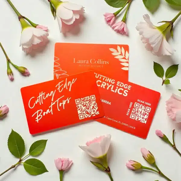 Three vibrant orange .8mm Engraved Aluminum Business Cards with white text and QR codes are displayed among pink flowers and green leaves on a white surface. Each card showcases unique designs and business details.