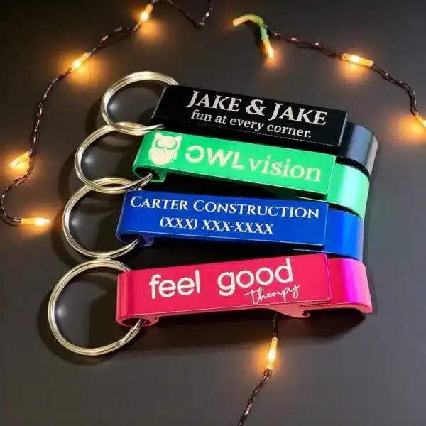 Four Custom Engraved Bottle Opener Keychains with company names—JAKE & JAKE, OWL vision, CARTER CONSTRUCTION, and feel good therapy—are displayed beside a string of small lights on a dark surface.
