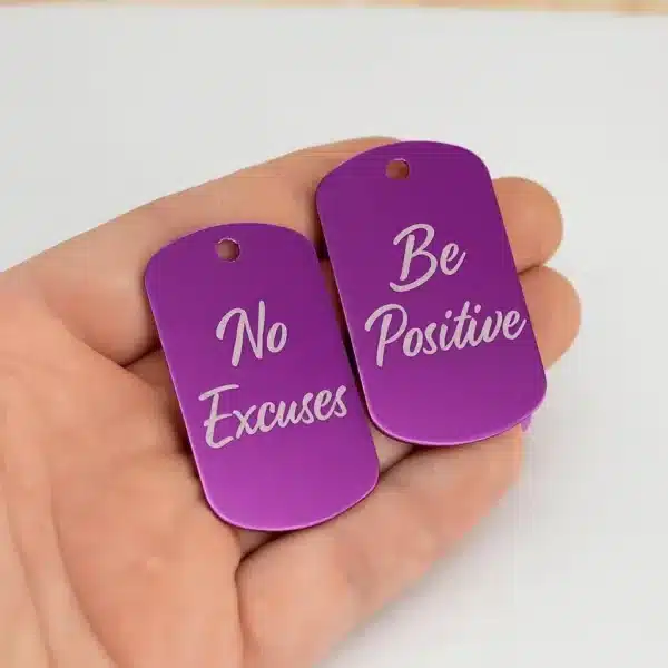A hand holds two Engraved Dog Tags with Optional Chain in purple—one reads "No Excuses" and the other "Be Positive," both in white script font.