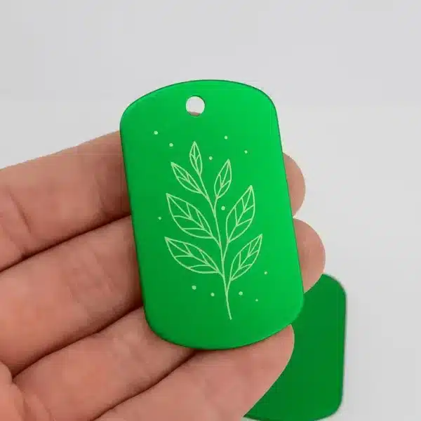 A hand holds the Engraved Dog Tag with Optional Chain, a green rectangular tag with rounded edges decorated with a white line drawing of a leafy branch and small dots.
