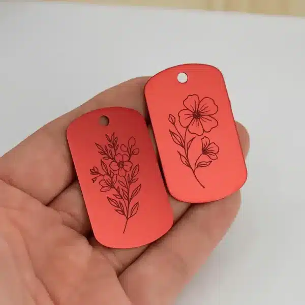 A hand holds two red Engraved Dog Tag with Optional Chain, each featuring a delicate floral design with blooming flowers and leaves. The tags have a hole at the top for easy attachment to a chain or keyring.