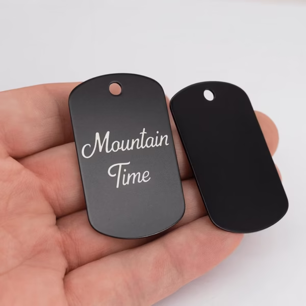 A hand holds two Engraved Dog Tags with Optional Chain; one tag is blank, and the other features "Mountain Time" engraved in white script.