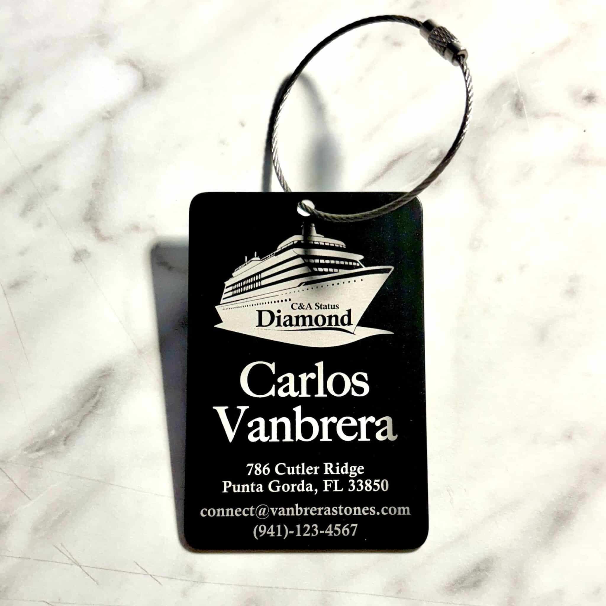 A Personalized Custom Engraved Metal Luggage Tag, black with a cruise ship graphic, “C&A Status Diamond,” Carlos Vanbrera’s info, and chain, is shown on a white surface.