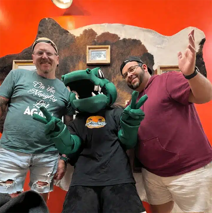 Two team members posing with a green alligator mascot inside a custom engraving workshop