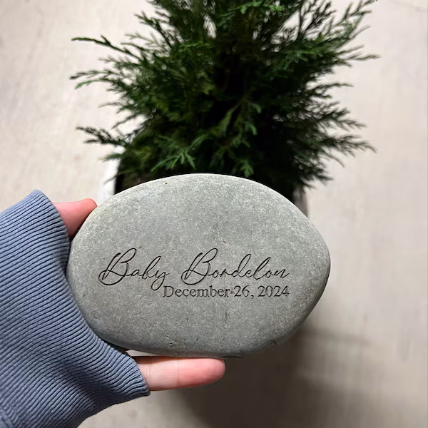 Hand holding a smooth personalized grey river stone engraved with the name Baby Banderas and the date December 26, 2024, shown in front of a small green plant.