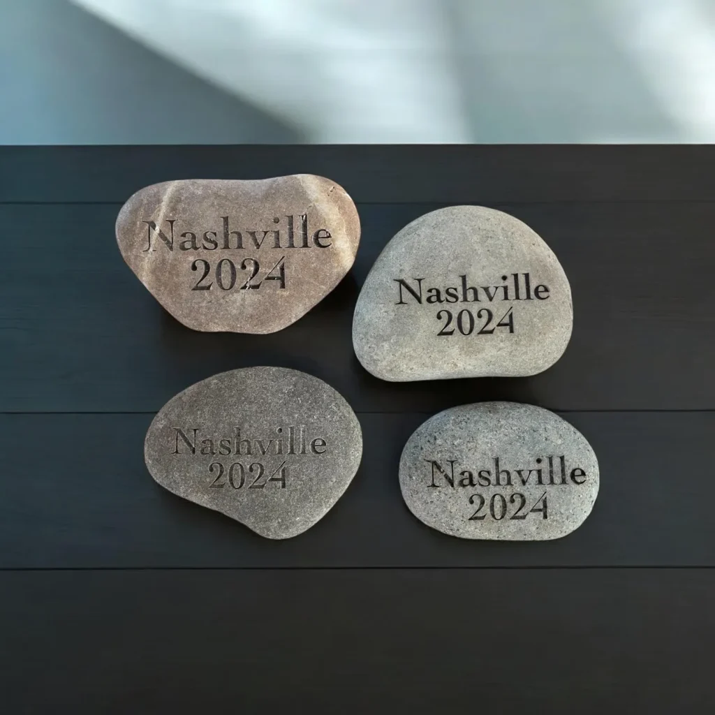 Four personalized engraved river stones of different shapes and shades displaying the text “Nashville 2024,” arranged on a dark wooden surface.