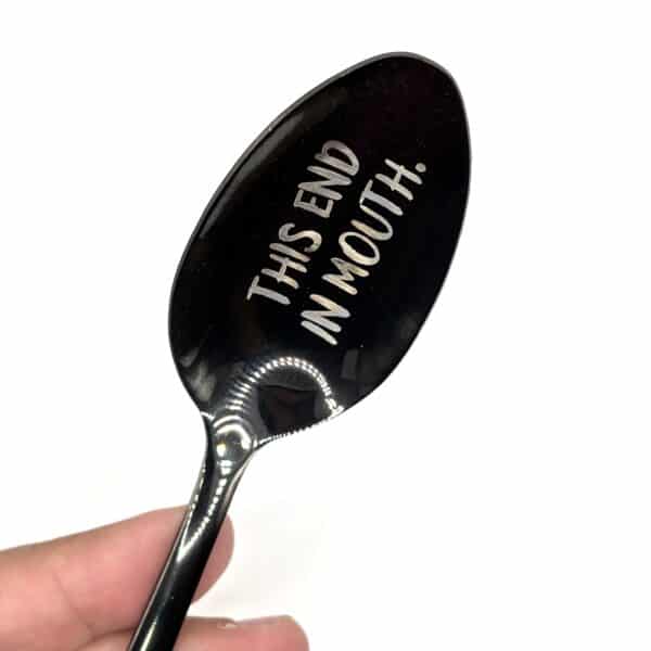 A hand holds the 8.75" Custom Black Engraved Spoon, featuring "THIS END IN MOUTH!" engraved on the bowl, set against a plain white background.