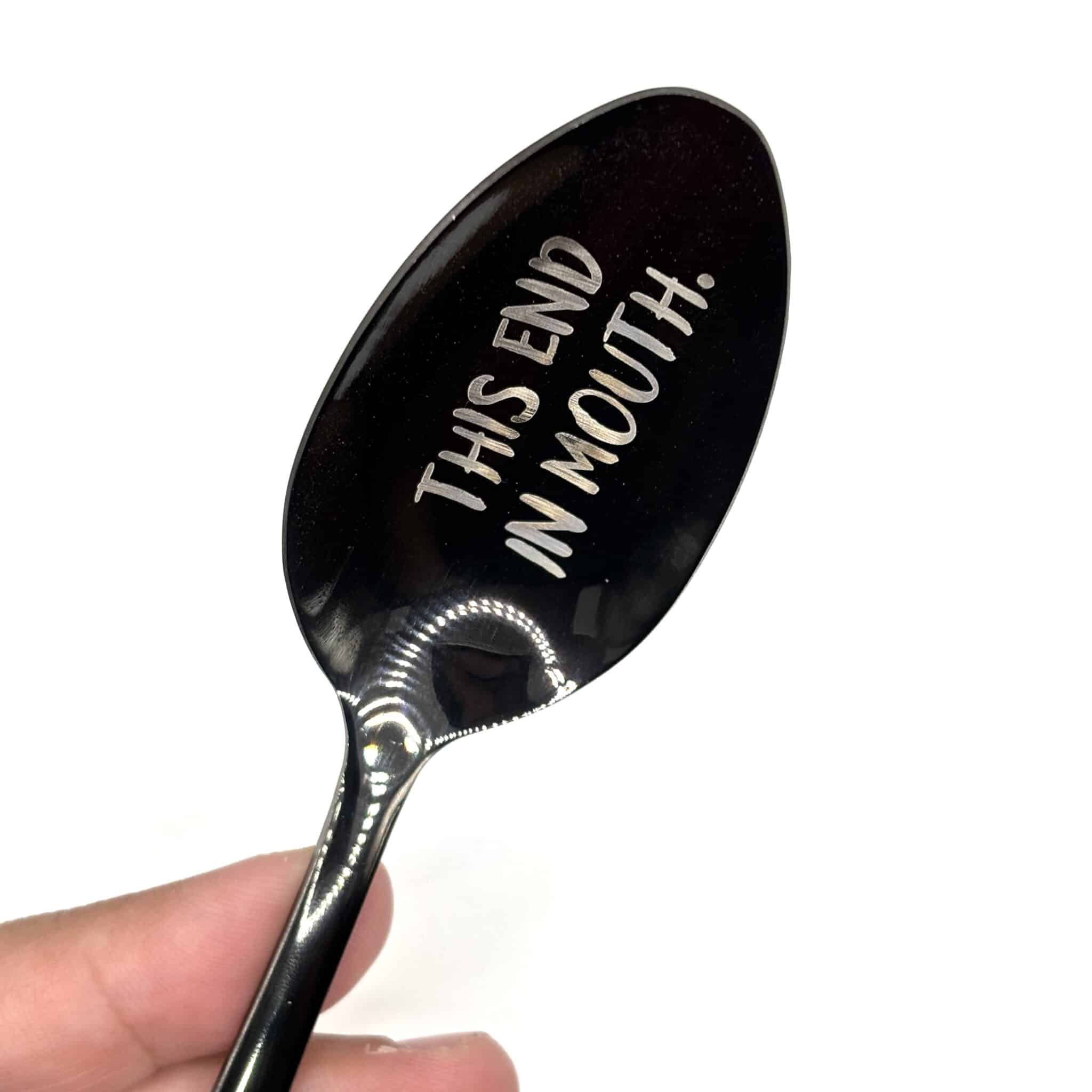 A hand holds the 8.75" Custom Black Engraved Spoon, featuring "THIS END IN MOUTH!" engraved on the bowl, set against a plain white background.