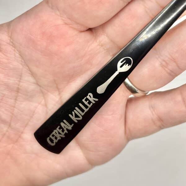 A hand holds the 8.75" Custom Black Engraved Spoon, featuring a spork image and "Cereal Killer" engraved on the handle.