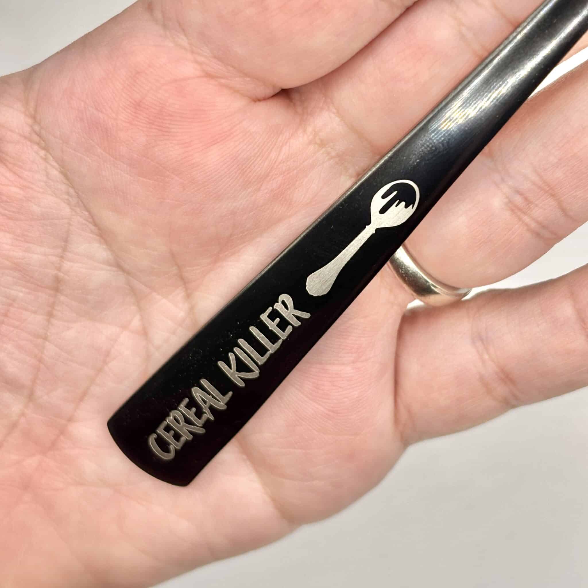 A hand holds the 8.75" Custom Black Engraved Spoon, featuring a spork image and "Cereal Killer" engraved on the handle.