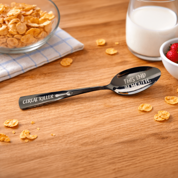 An 8.75" Custom Black Engraved Spoon labeled "Cereal Killer" and "This End In Mouth" rests on a wooden table near cereal flakes, a bowl of cornflakes, a glass of milk, and a dish of strawberries.