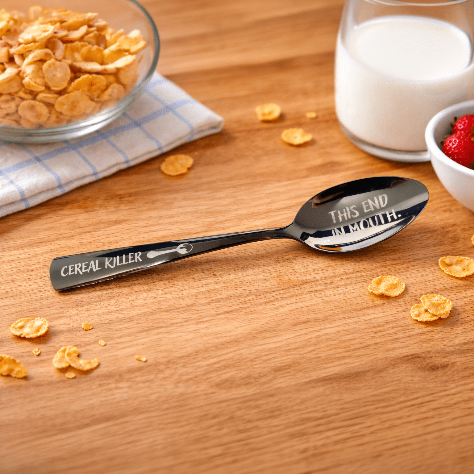 An 8.75" Custom Black Engraved Spoon labeled "Cereal Killer" and "This End In Mouth" rests on a wooden table near cereal flakes, a bowl of cornflakes, a glass of milk, and a dish of strawberries.