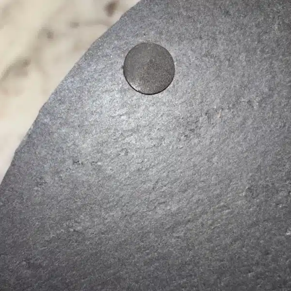 Close-up of a small round magnet attached to the edge of a dark, textured circular surface, possibly a slate board, with a blurred white and gray background.